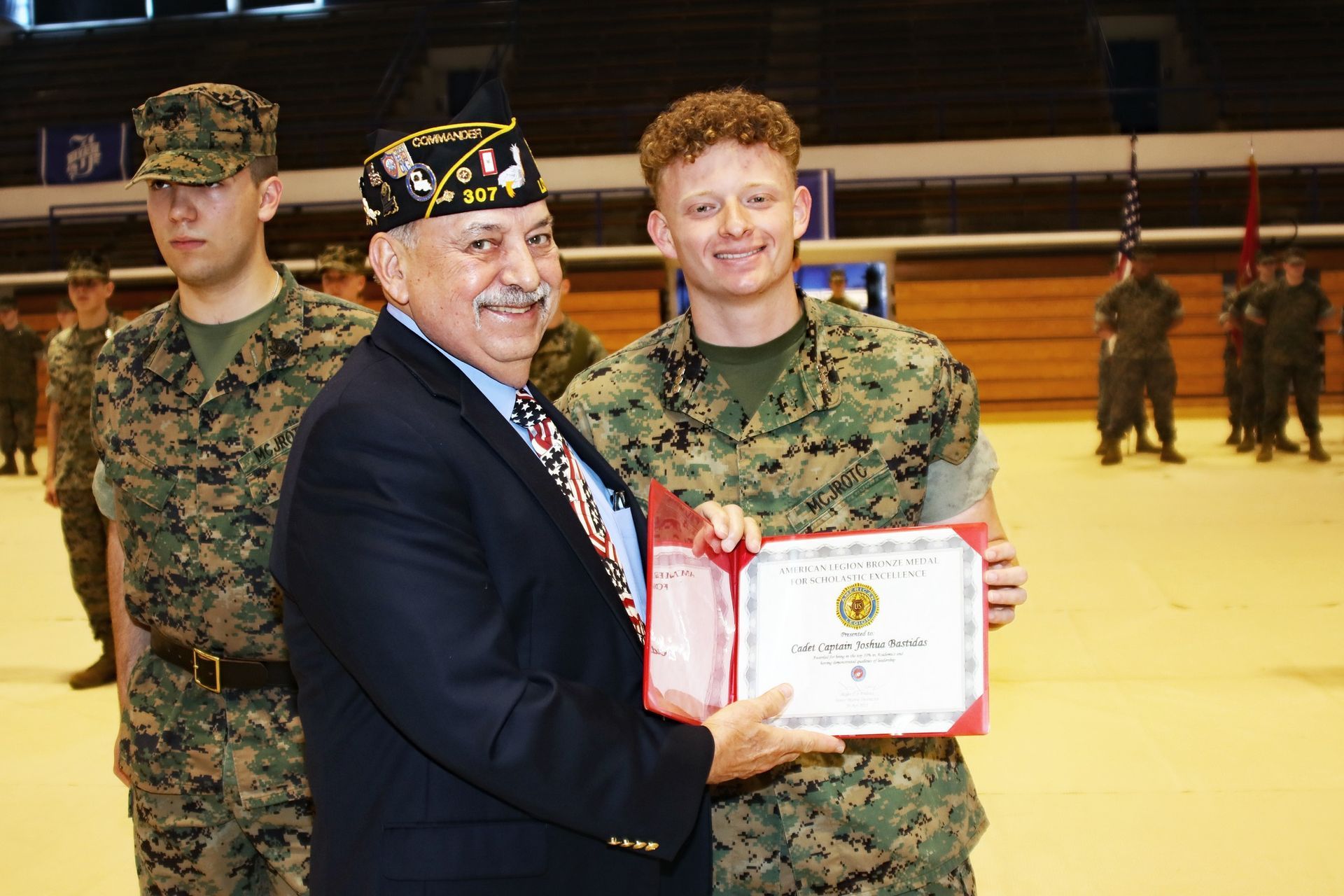 Military award presentation with smiling soldier holding a certificate beside a man in a suit and medals