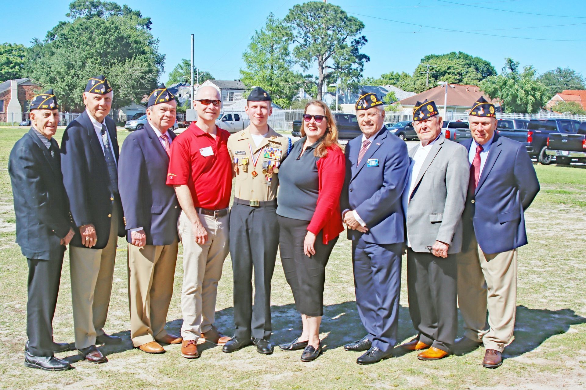 Group of people standing outdoors in front of parked cars, posing in bright sunlight.