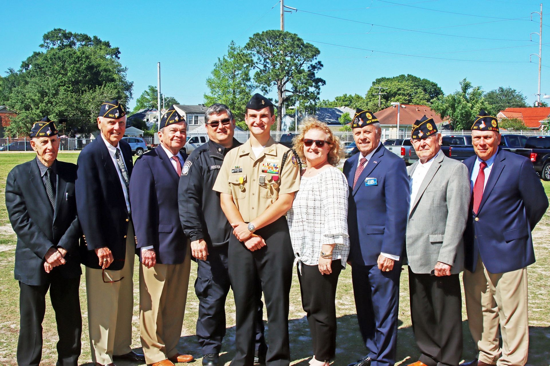 Group of officials posing outdoors on a sunny day, with trees and buildings in the background.