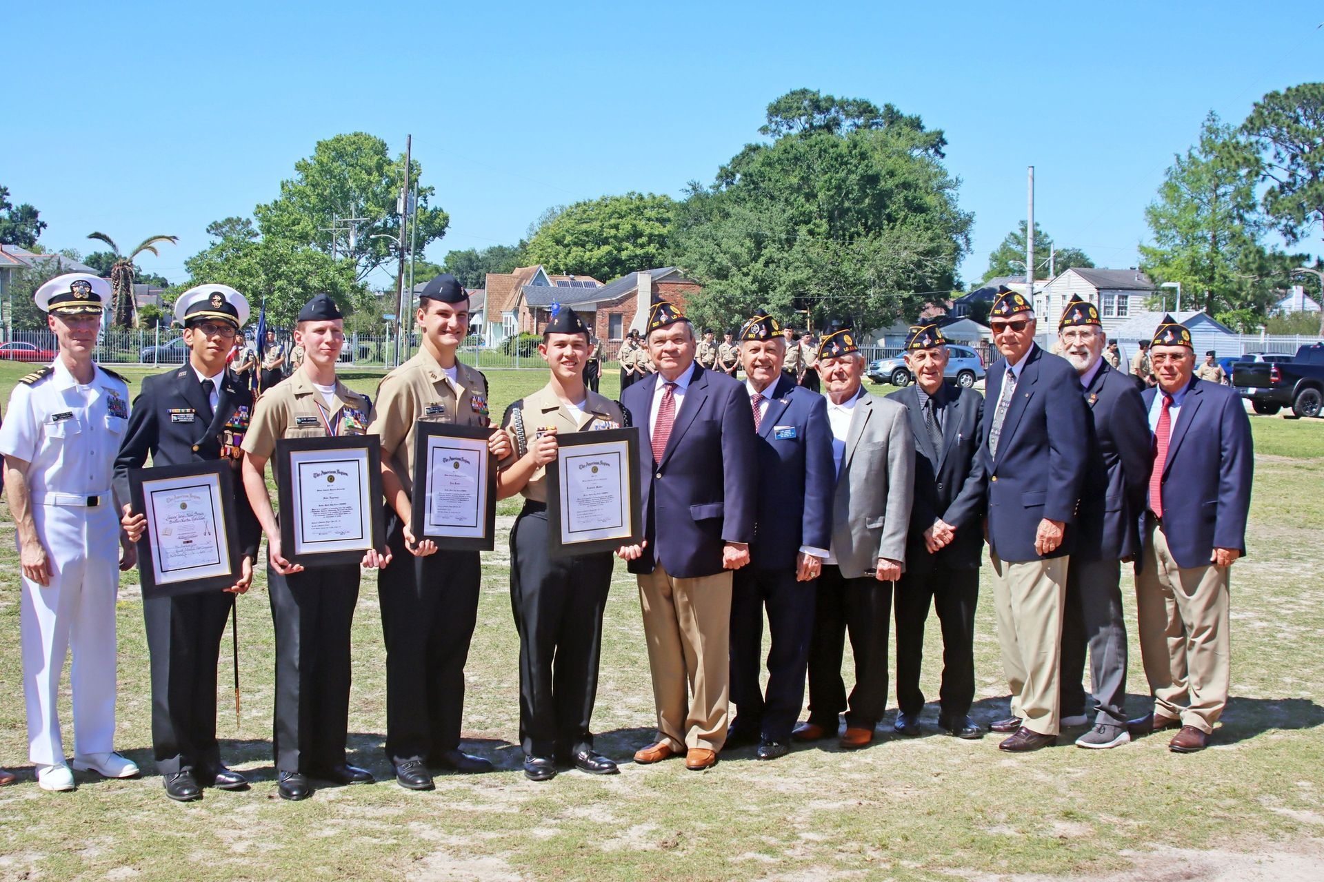 Group of uniformed and suited people posing outdoors, several holding framed certificates during an awards ceremony.