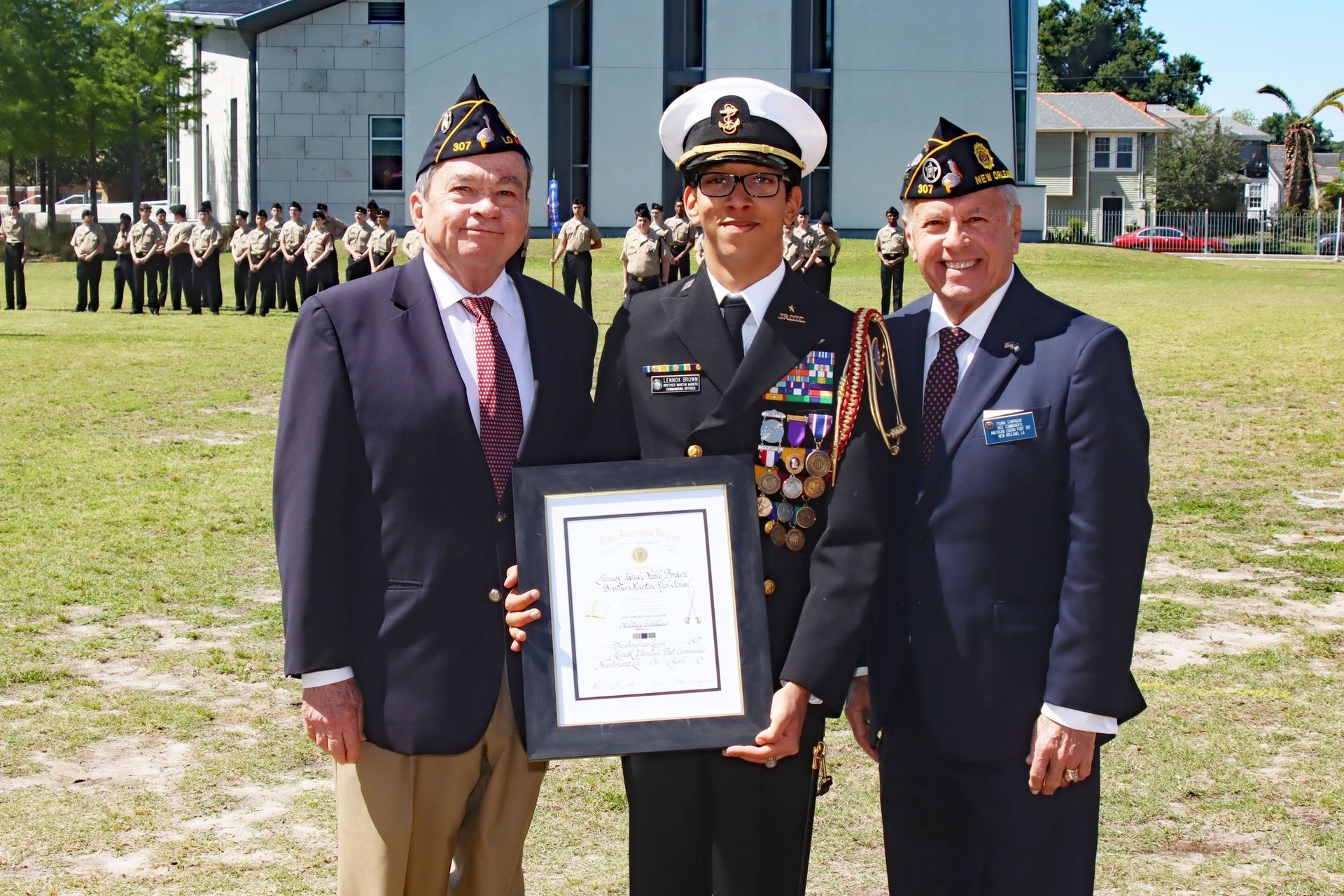 Three men in formal attire stand outdoors; the center man in uniform holds a framed certificate.