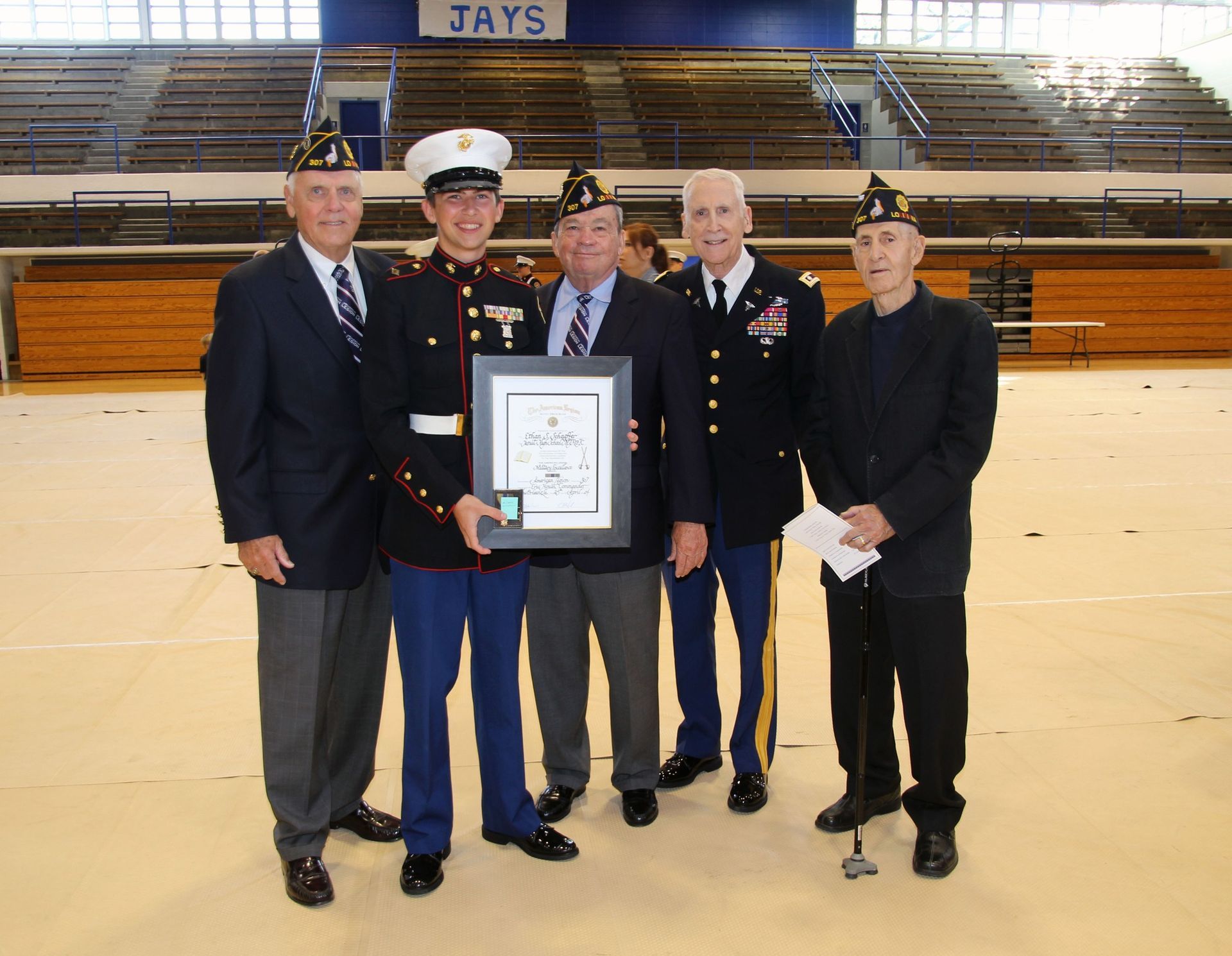 Five men on a gym floor holding a framed certificate, with bleachers in the background