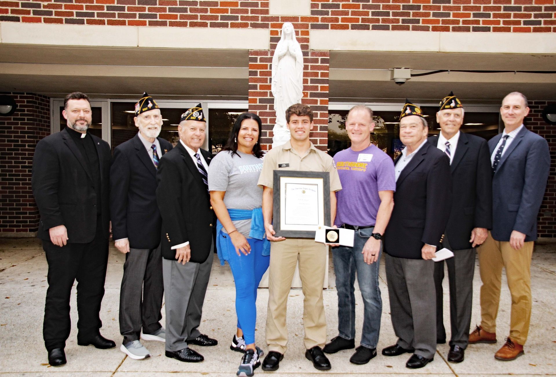 Group posing outdoors in front of a brick building, one person holding a certificate plaque.