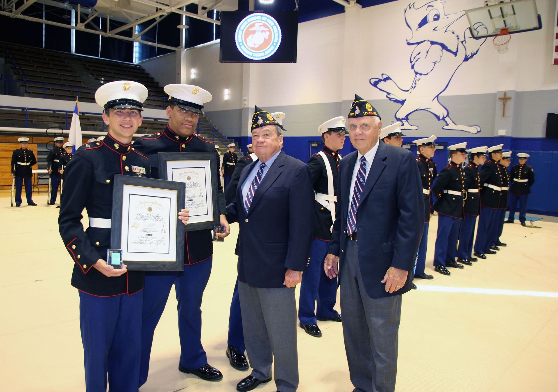 Two uniformed cadets hold framed awards beside a suited man in a gymnasium during a ceremony