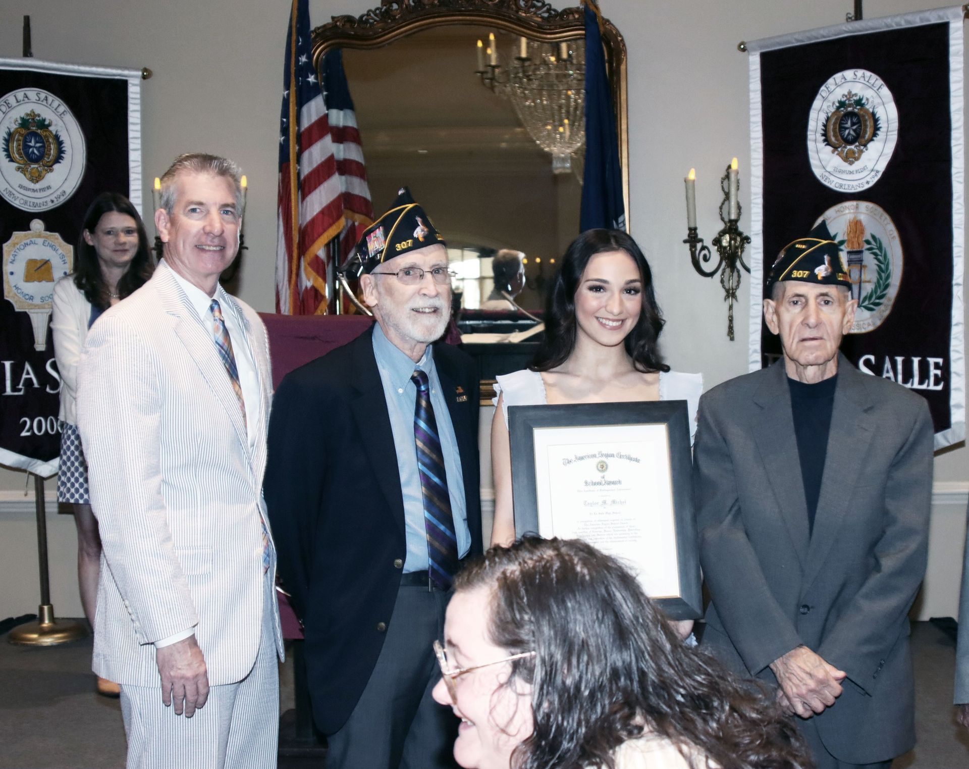 Group of people posing at a formal ceremony, one woman holding a framed certificate in front of banners and flags.