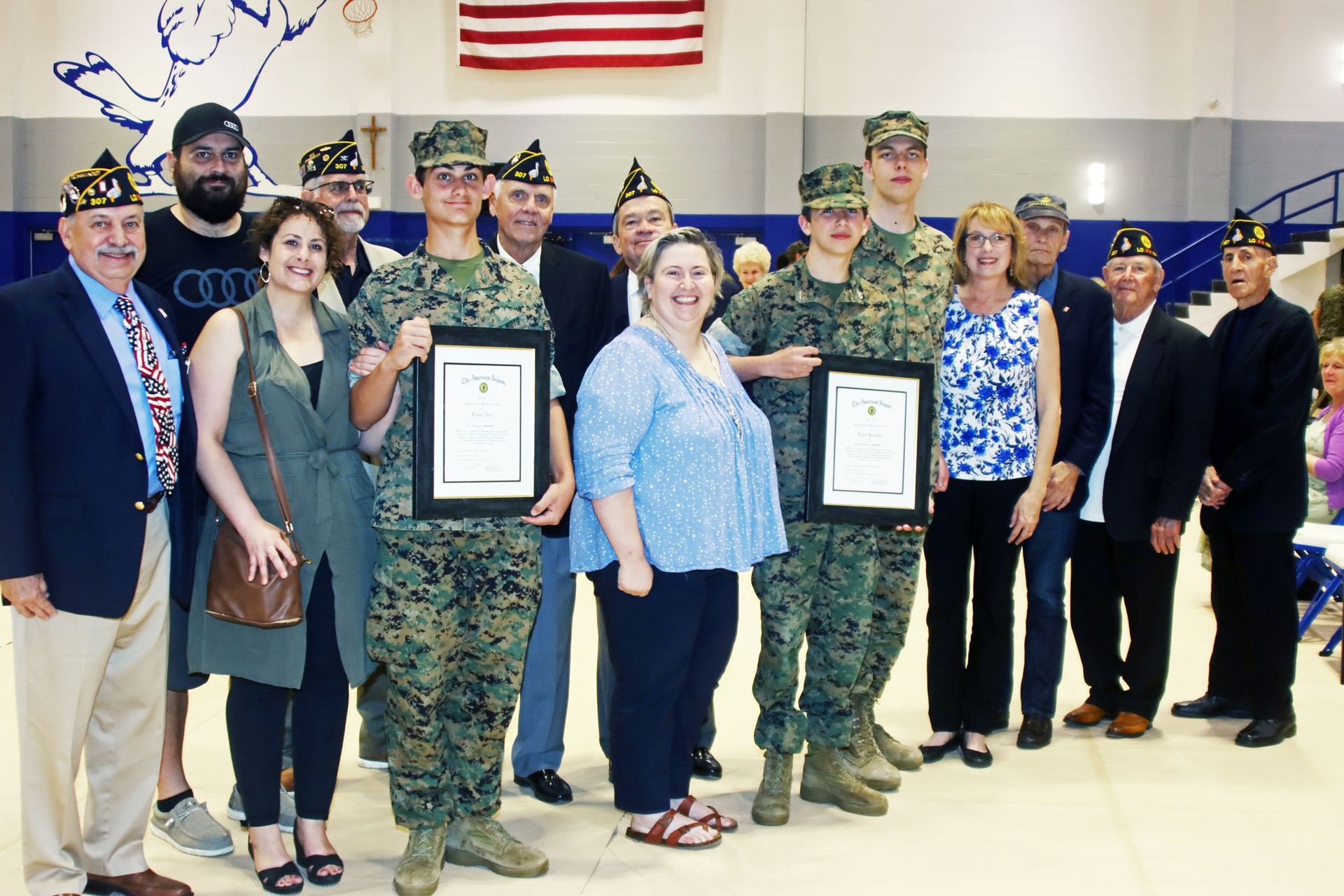 Group of uniformed military personnel and civilians posing indoors with framed certificates and an American flag above them