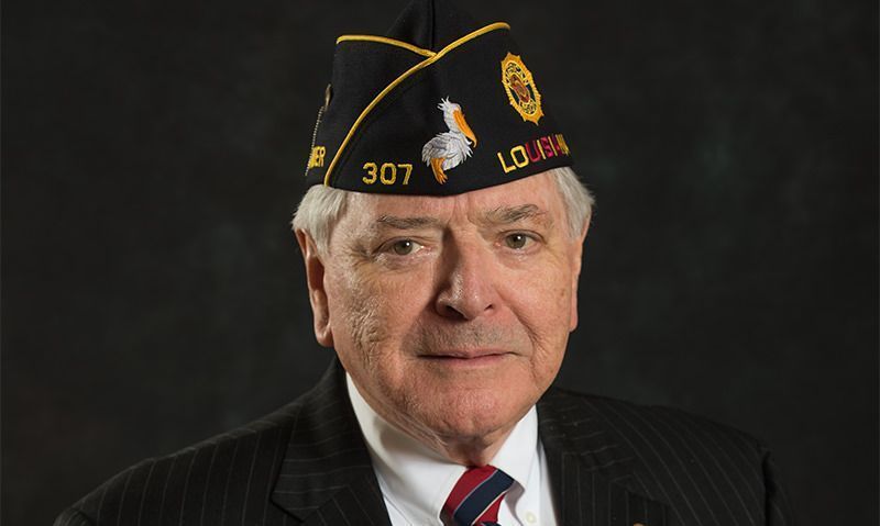 Portrait of a man in a dark suit and military veteran cap against a black background.