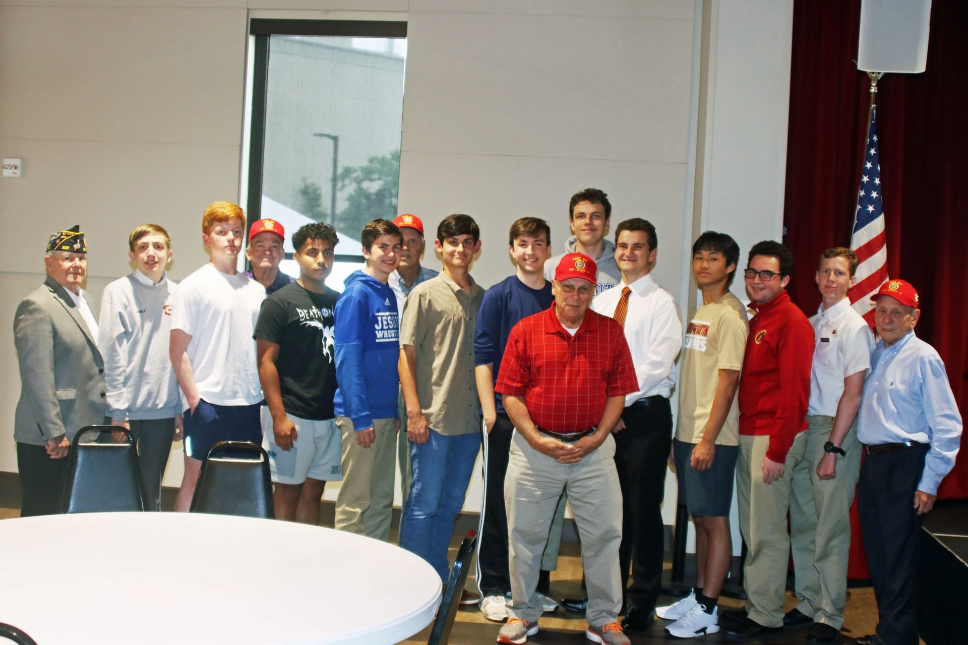 Group of people posing indoors beside an American flag, with one man in a red shirt centered in front.