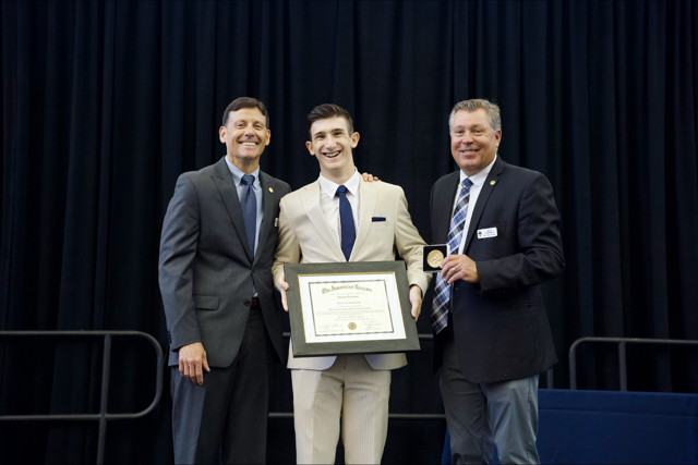 Three men onstage, one holding a framed certificate, posing in front of a black curtain.