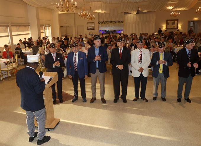 People standing in a banquet hall, each holding a drink, while a speaker addresses them from a podium.