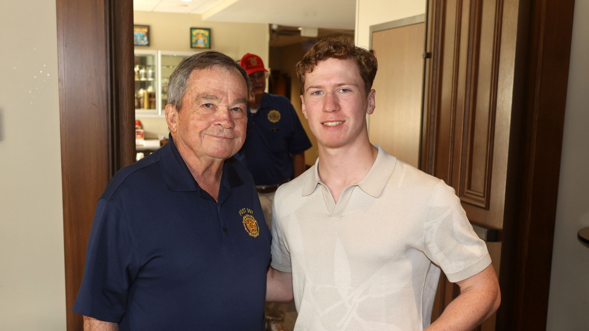 Two men posing indoors, one in a navy polo and one in a white shirt, standing in a doorway.
