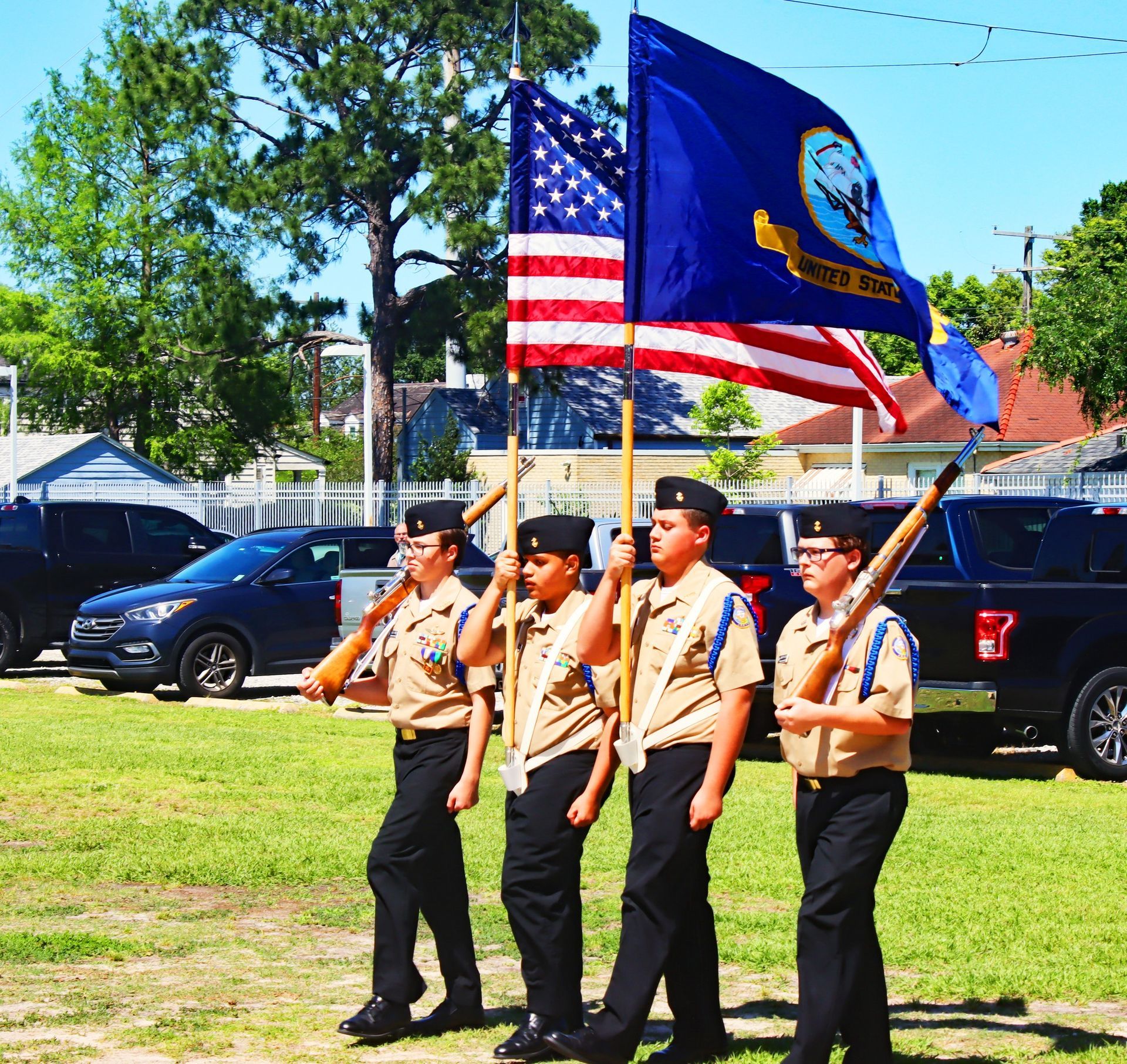 Color guard in tan uniforms marches carrying the U.S. and New Mexico flags on a grassy field.