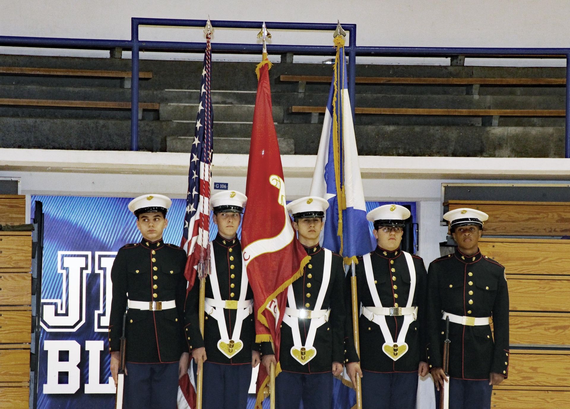 Four uniformed cadets stand with flags on a gymnasium stage