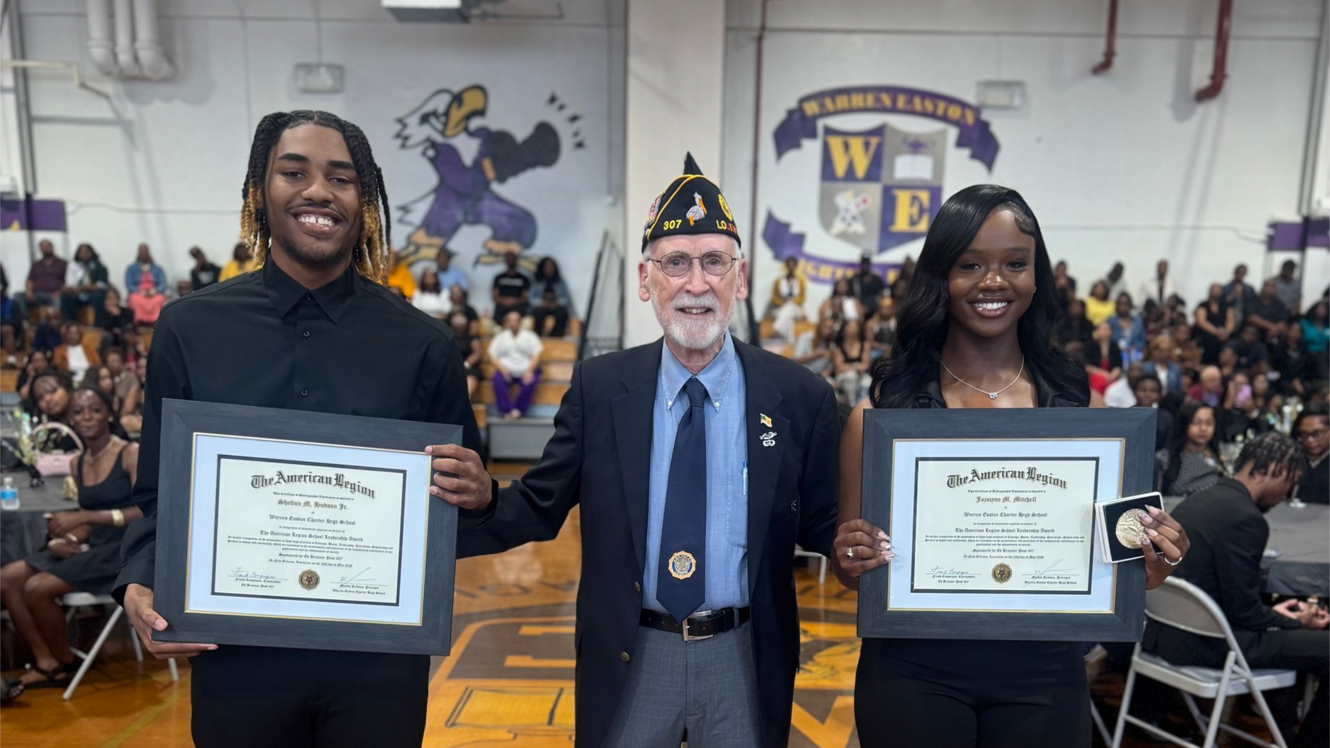 Two people hold framed certificates beside a man in uniform at a school awards ceremony.