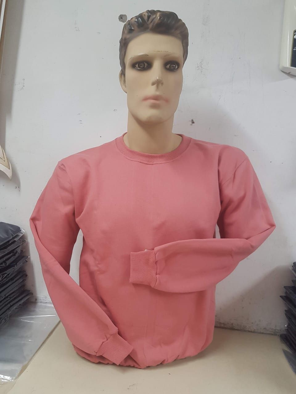 Un maniquí con una sudadera rosa lisa de manga larga y cuello redondo, colocado frente a una pared de color neutro.