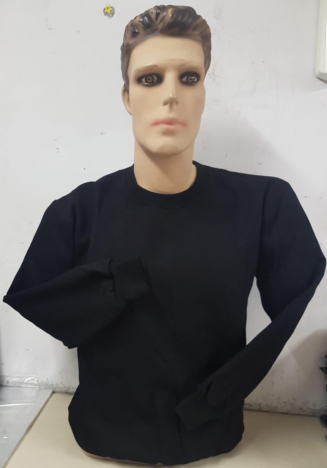 Un torso de maniquí masculino con un suéter negro liso de manga larga y cuello redondo sobre un fondo liso de color claro.