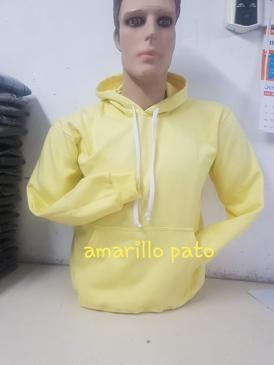 Una sudadera amarilla con capucha y cordones blancos, expuesta sobre el torso de un maniquí contra una pared blanca.