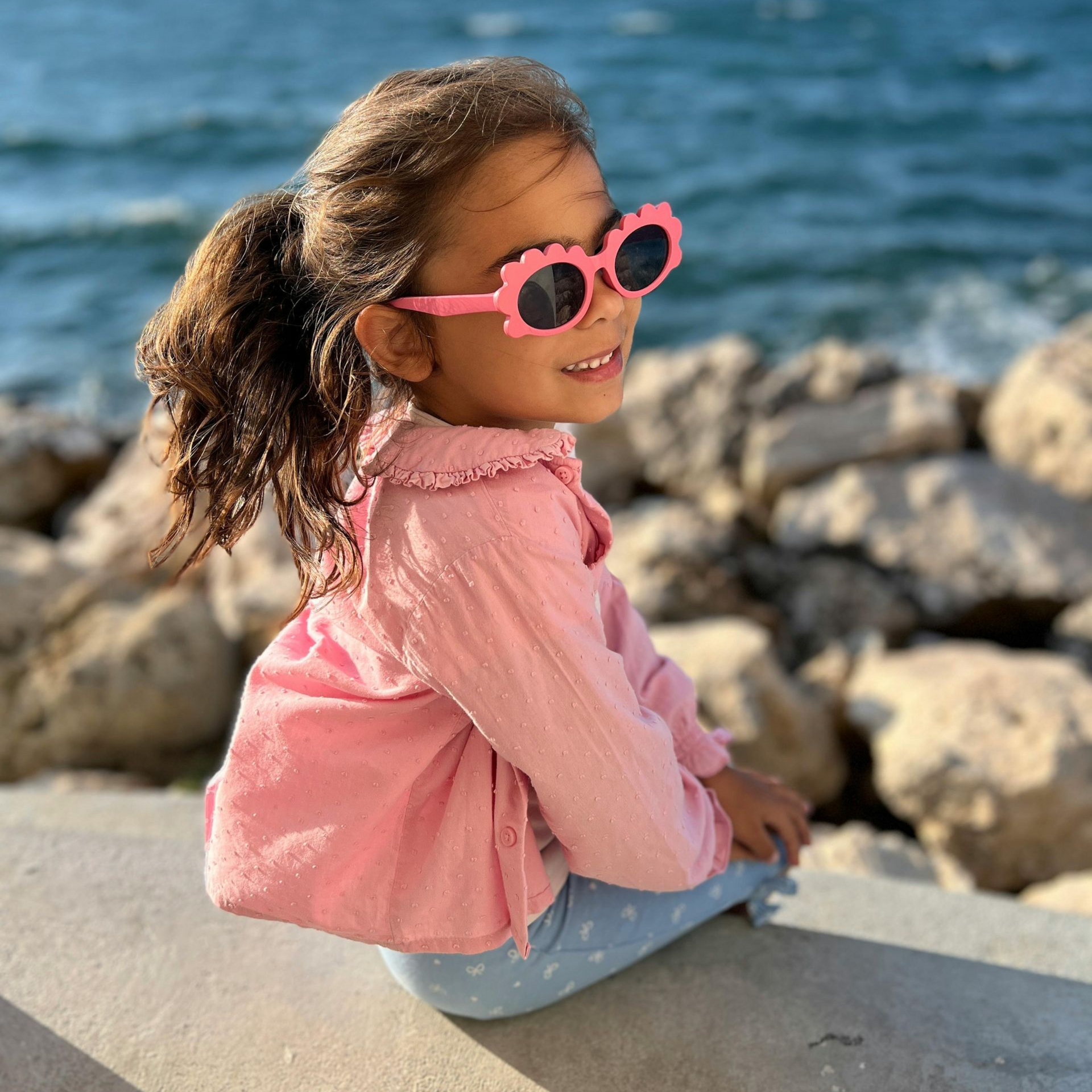 Un niño con gafas de sol rosas con bordes ondulados y una camiseta rosa está sentado en una cornisa con vistas al océano.