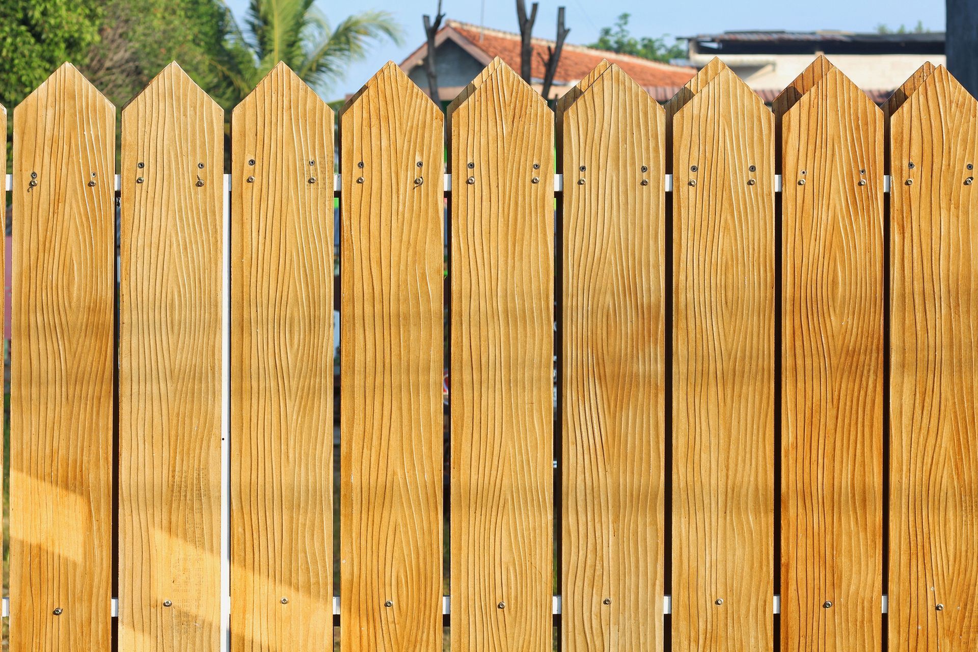 Wooden fence with pointed tops, outdoors, with a house and trees in the background.