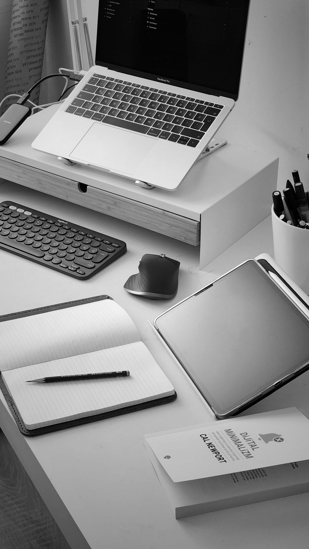 Postazione di lavoro in bianco e nero con laptop su supporto, tastiera, mouse, quaderno, penna e portapenne su una scrivania.