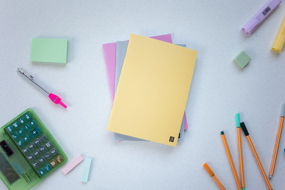 Un quaderno giallo con calcolatrice, matite, post-it e una chiavetta USB su una scrivania.