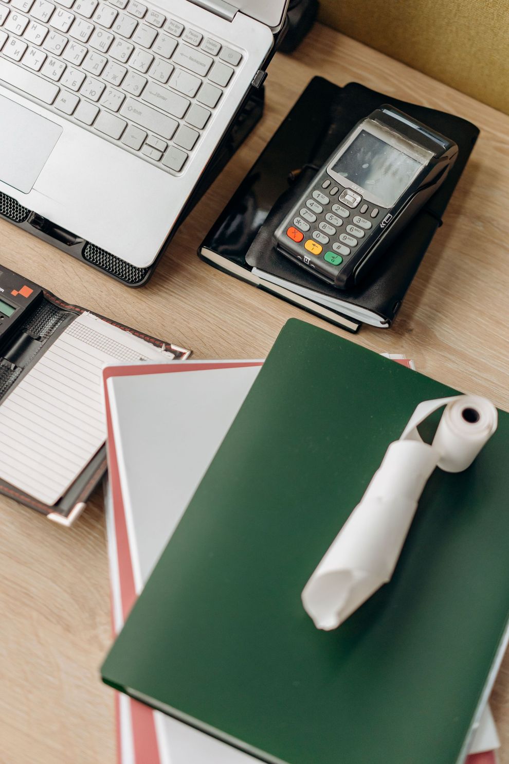 Scrivania con computer portatile, terminale di pagamento, quaderni e una penna su una cartella verde.