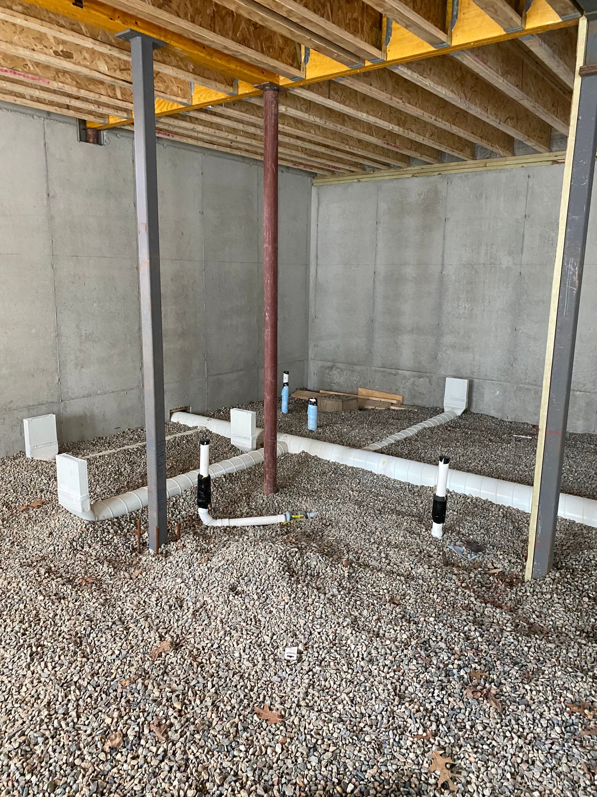 Under-construction basement with plumbing pipes, support beams, gravel floor, and exposed ceiling joists.