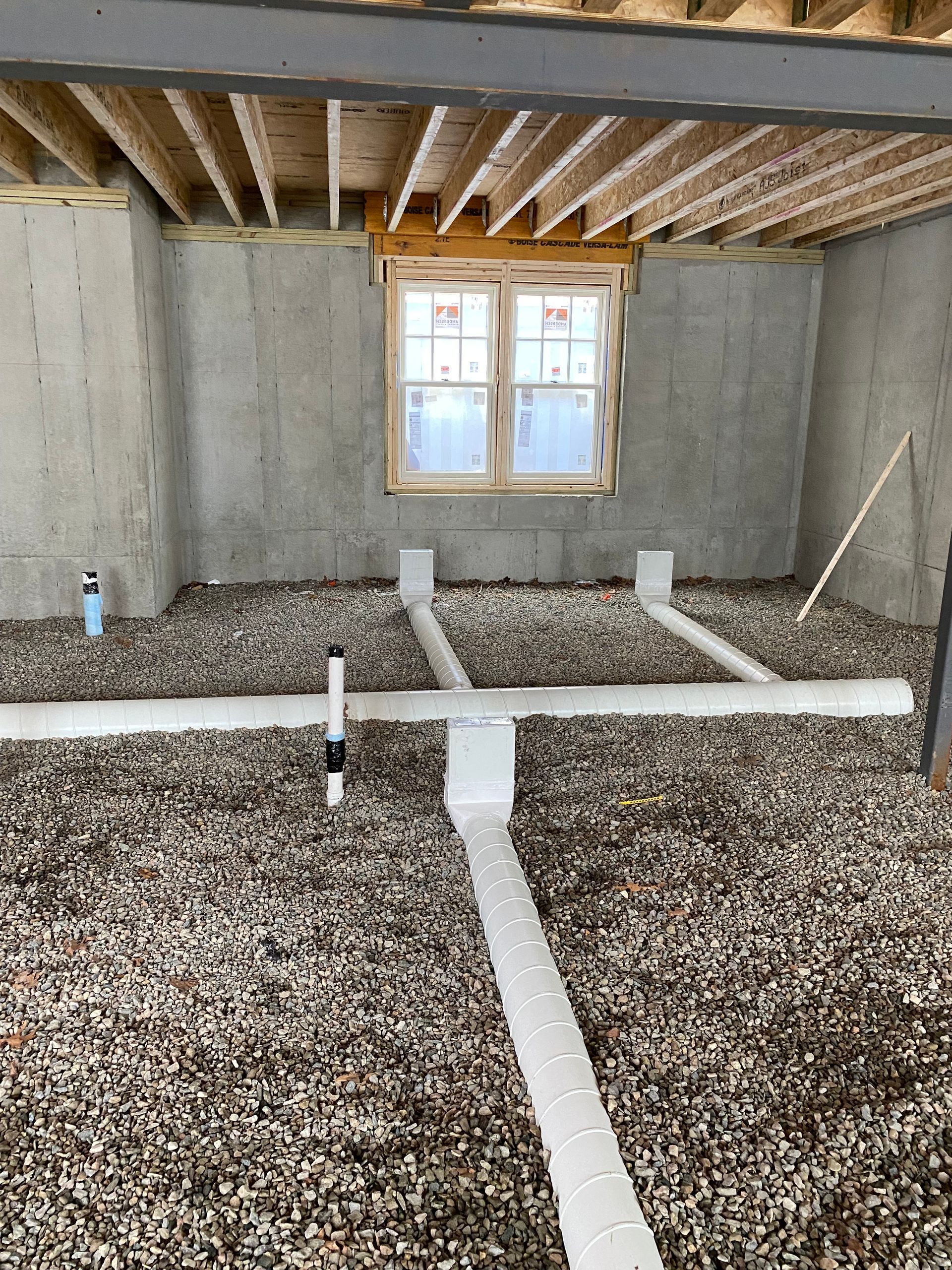 Basement construction showing drainage pipes laid in gravel, below a window, against concrete walls. Wooden beams overhead.