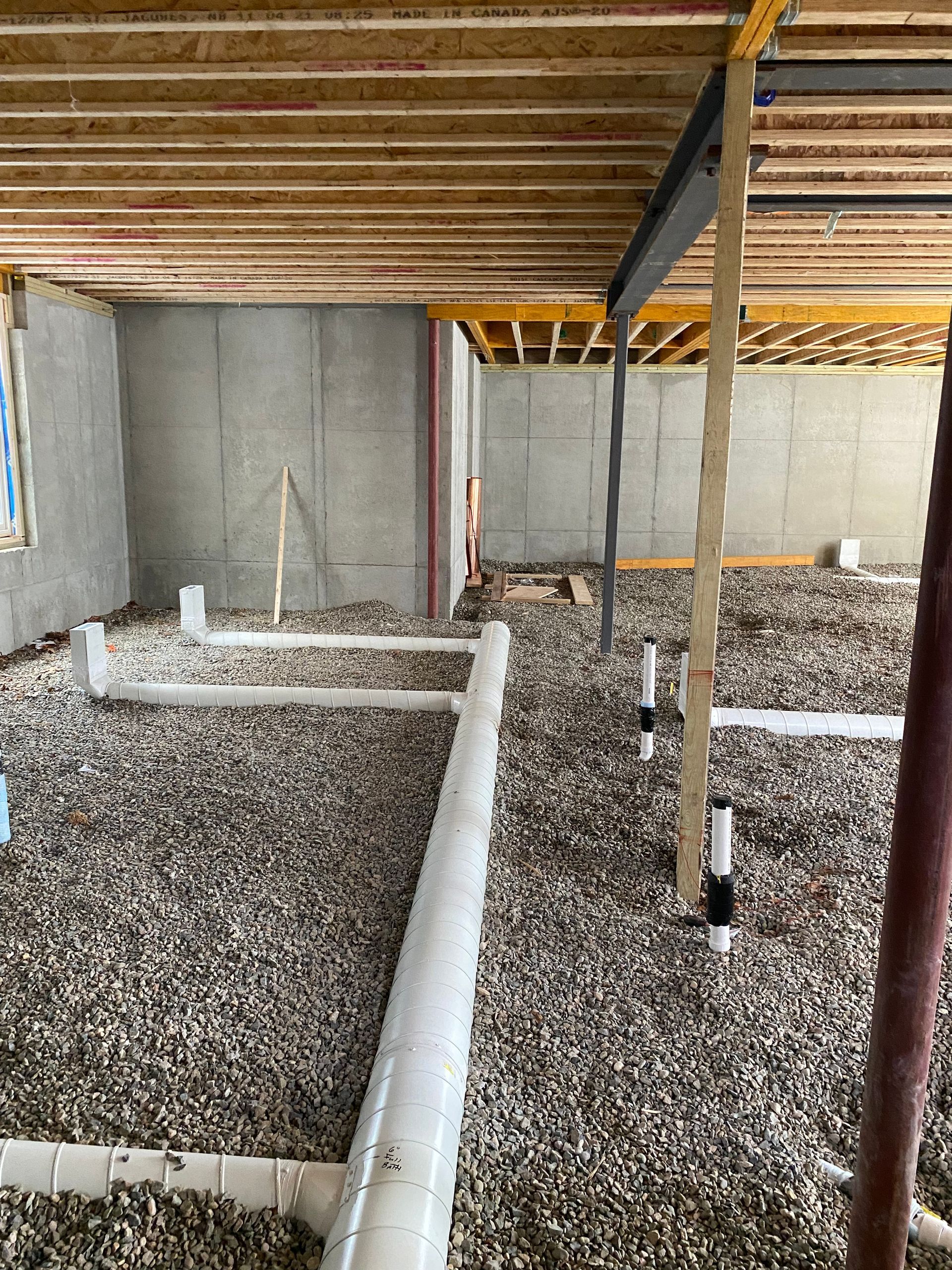 Basement under construction. Gray gravel floor, white pipes, wooden ceiling joists, and concrete walls.