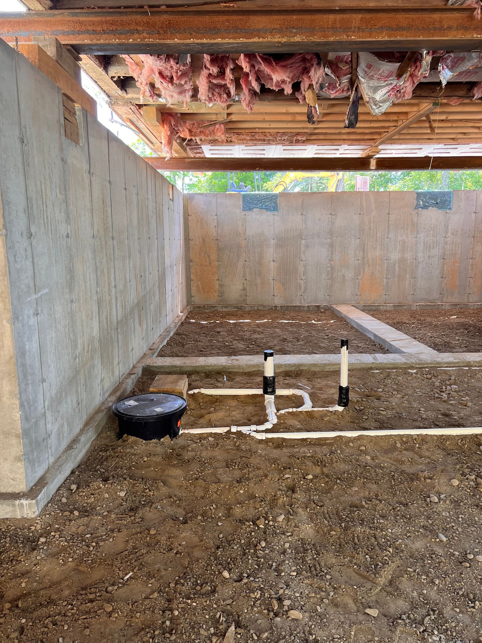 Concrete foundation with exposed pipes, dirt floor, and insulation under a wooden structure.