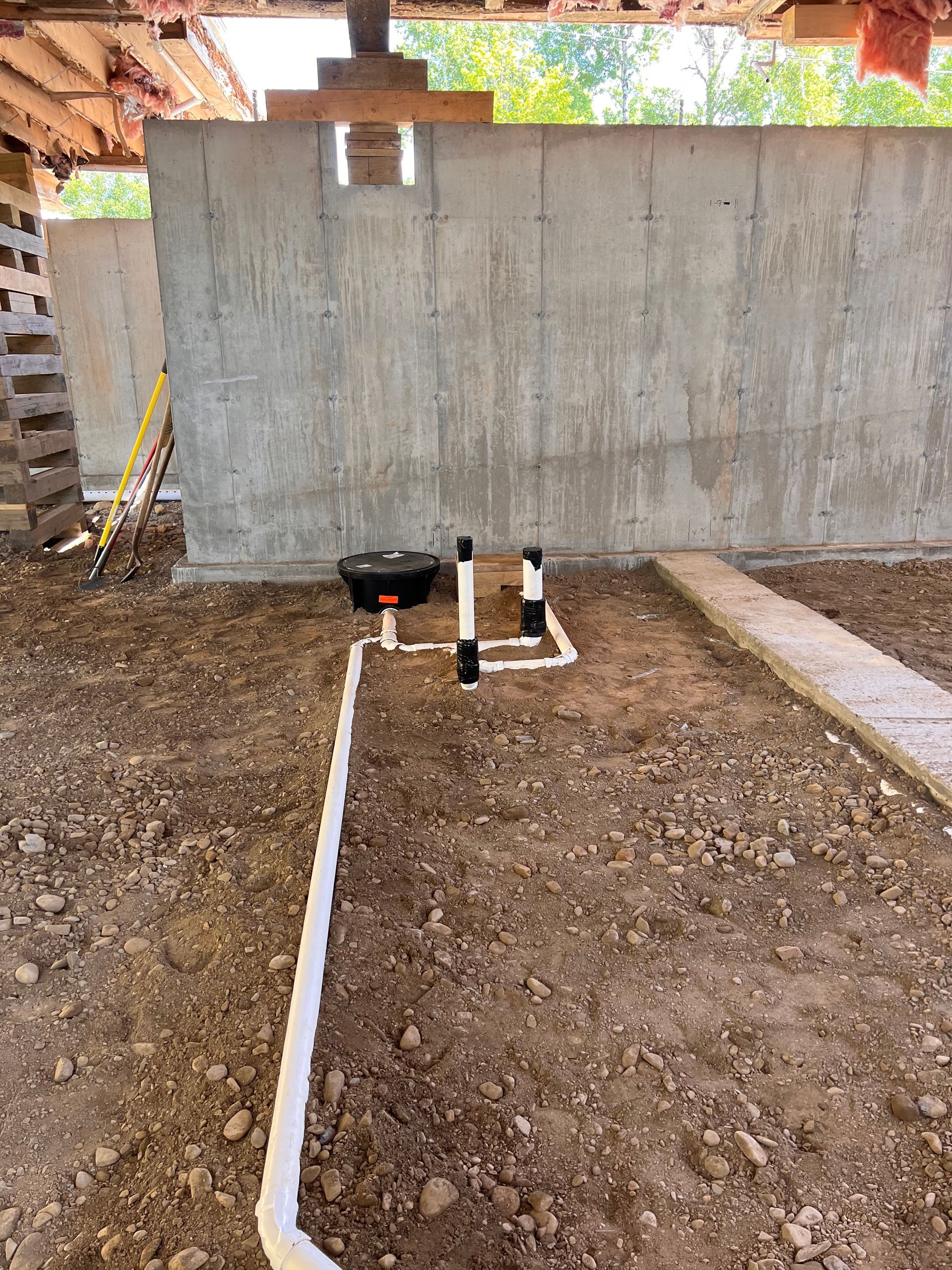 Plumbing pipes on dirt ground near a concrete wall; supports and beams overhead.
