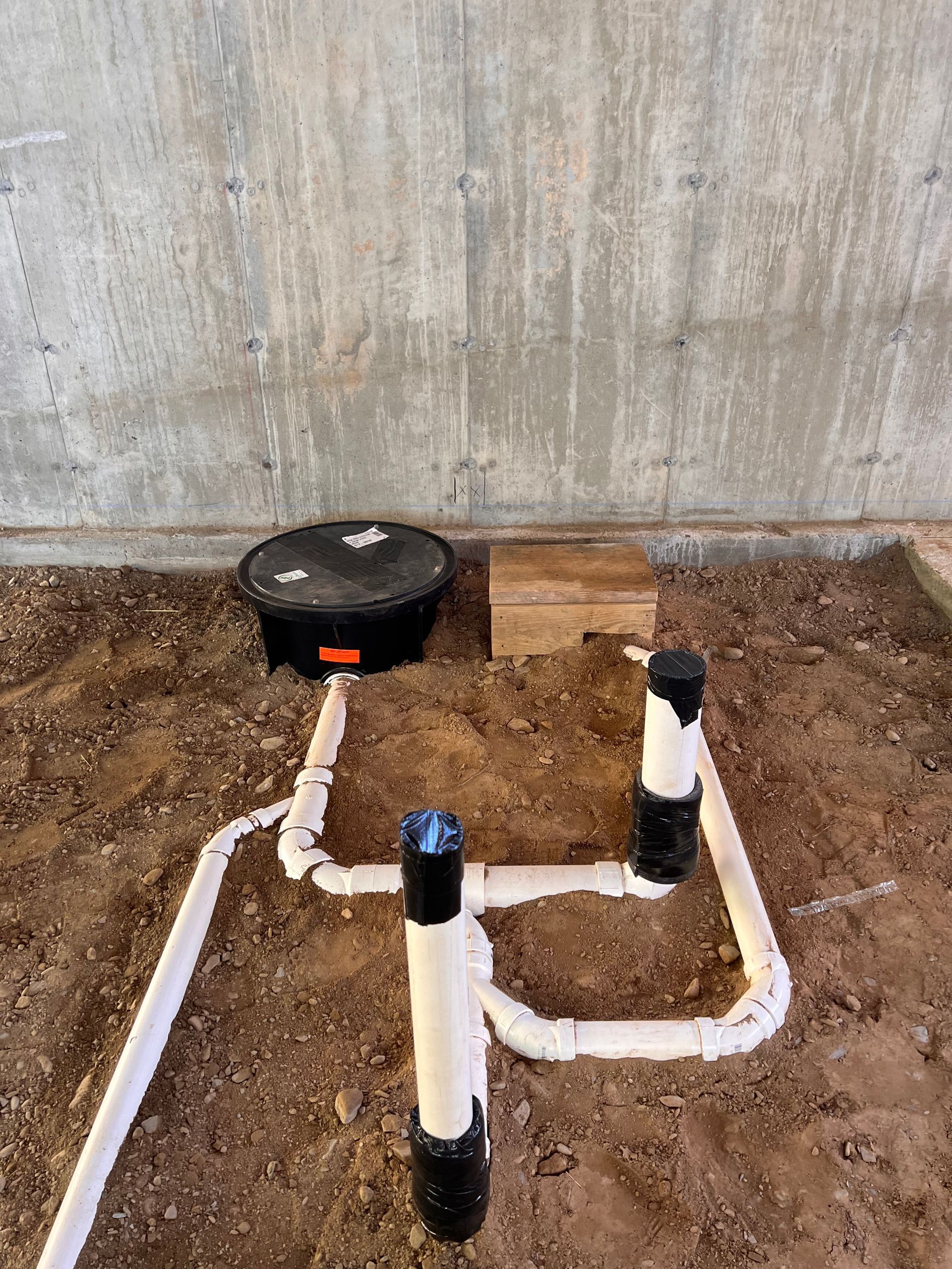Plumbing pipes laid in dirt, with a black container and a wooden block against a concrete wall.