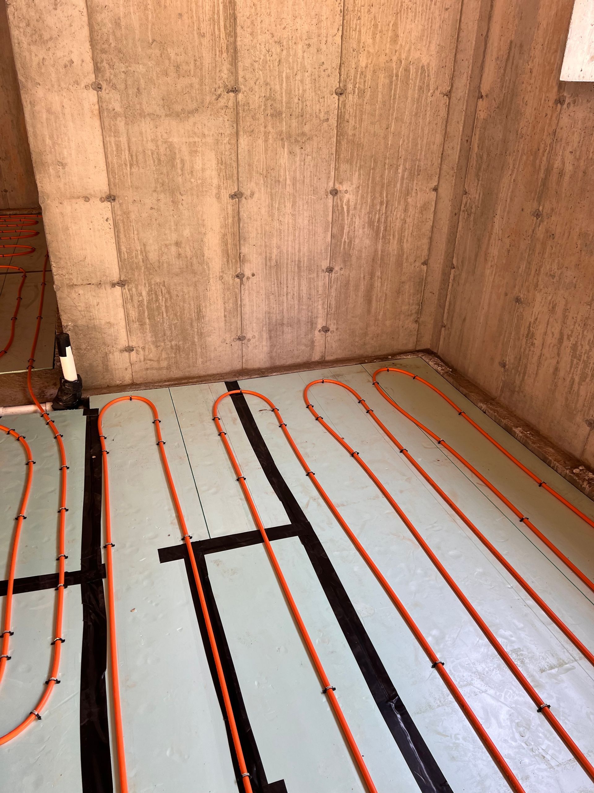 Orange radiant floor heating tubing laid on a light green surface, against a concrete wall.