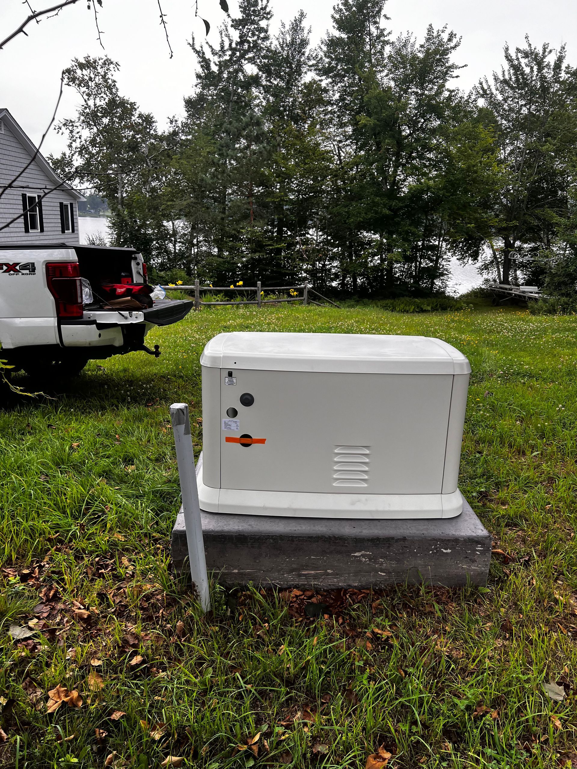 Gray and white standby generator on a concrete pad in a grassy yard, with a white truck in the background.