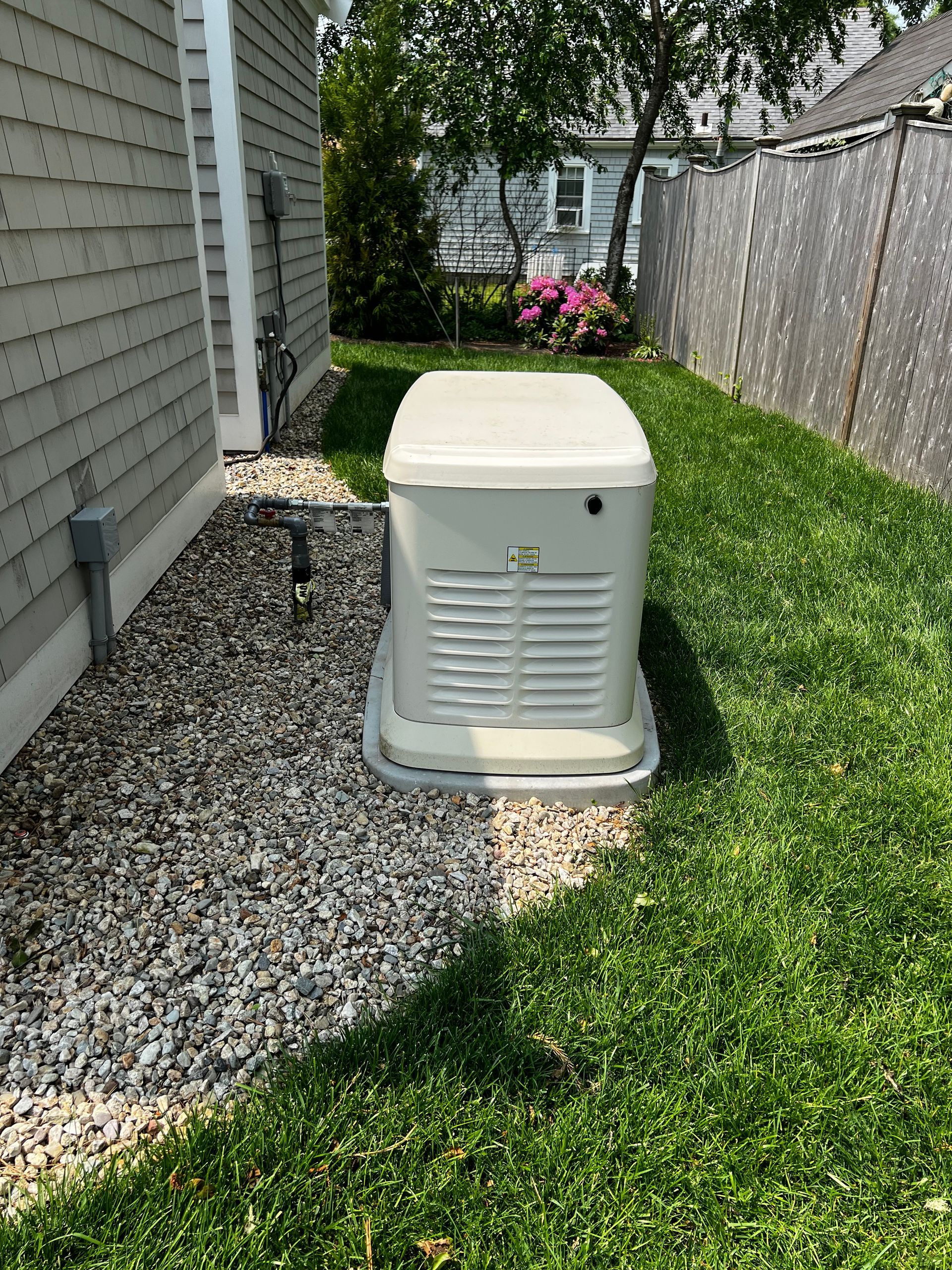 A beige standby generator sits on a gravel base next to a house with a wooden fence.