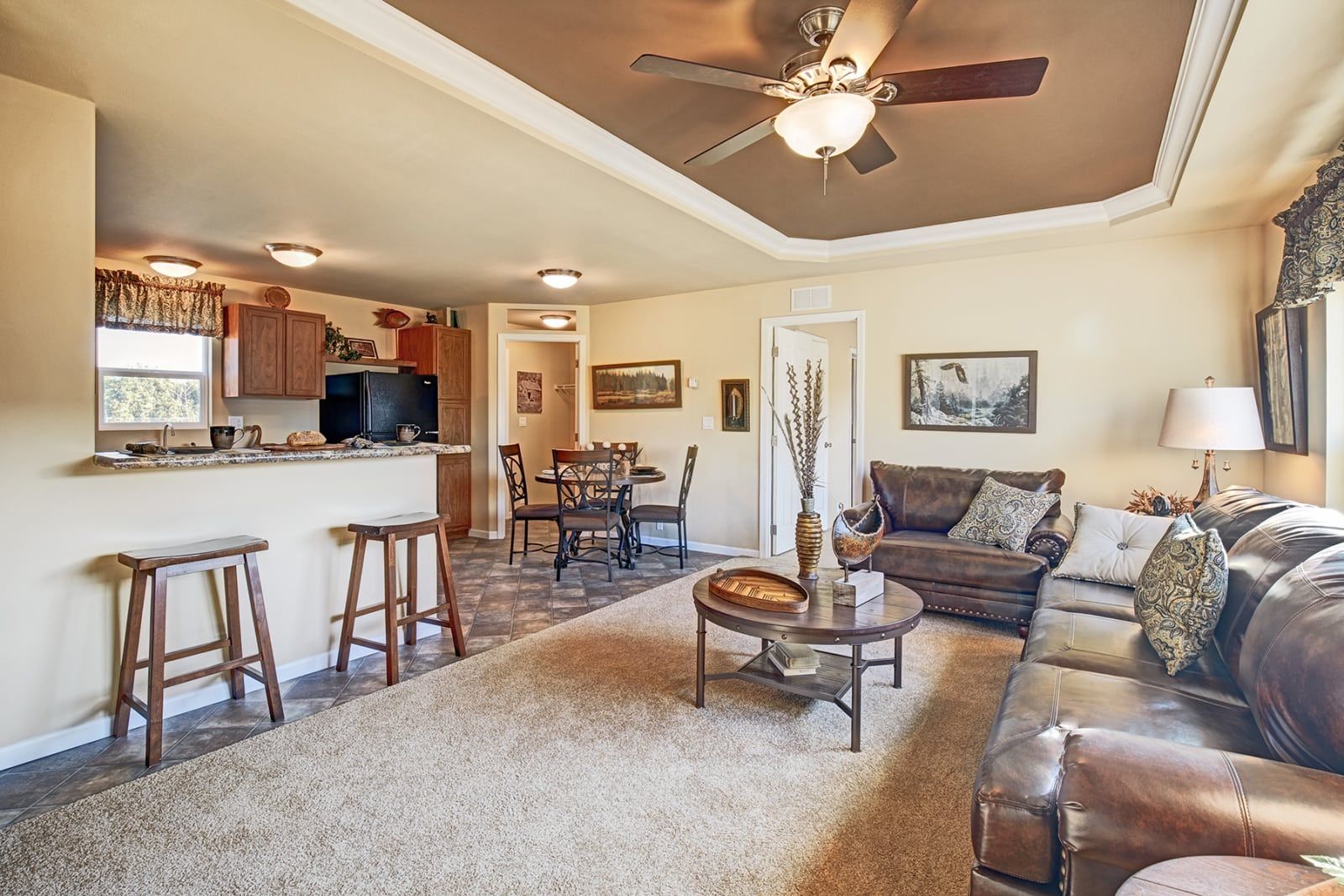 Open-concept living room with brown leather sofa, rug, and bar stools. Kitchen in the background.
