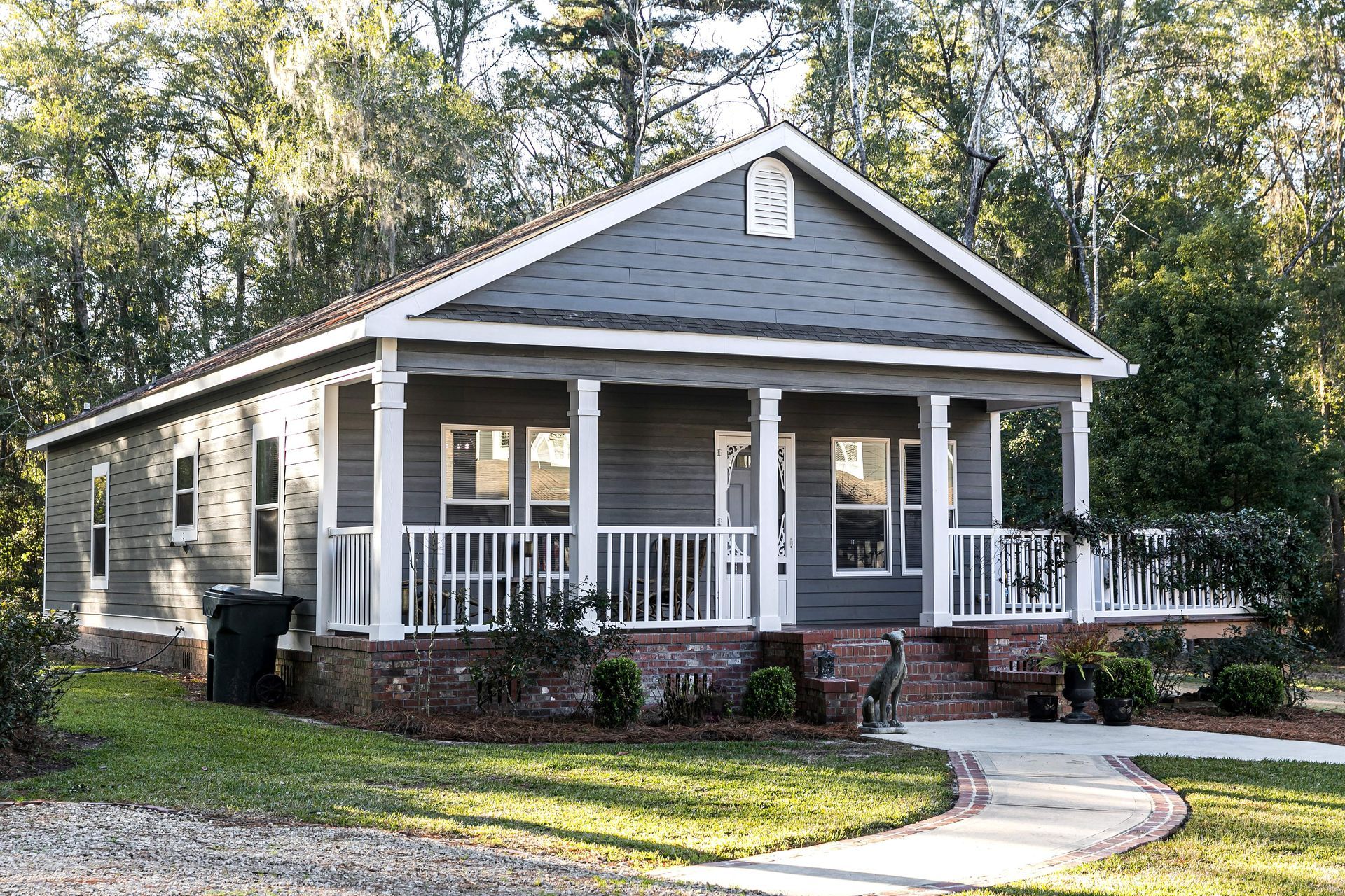 A manufactured home with a wide front porch sits on a landscaped lot surrounded by trees. A manufactured home with a wide front porch sits on a landscaped lot surrounded by trees.