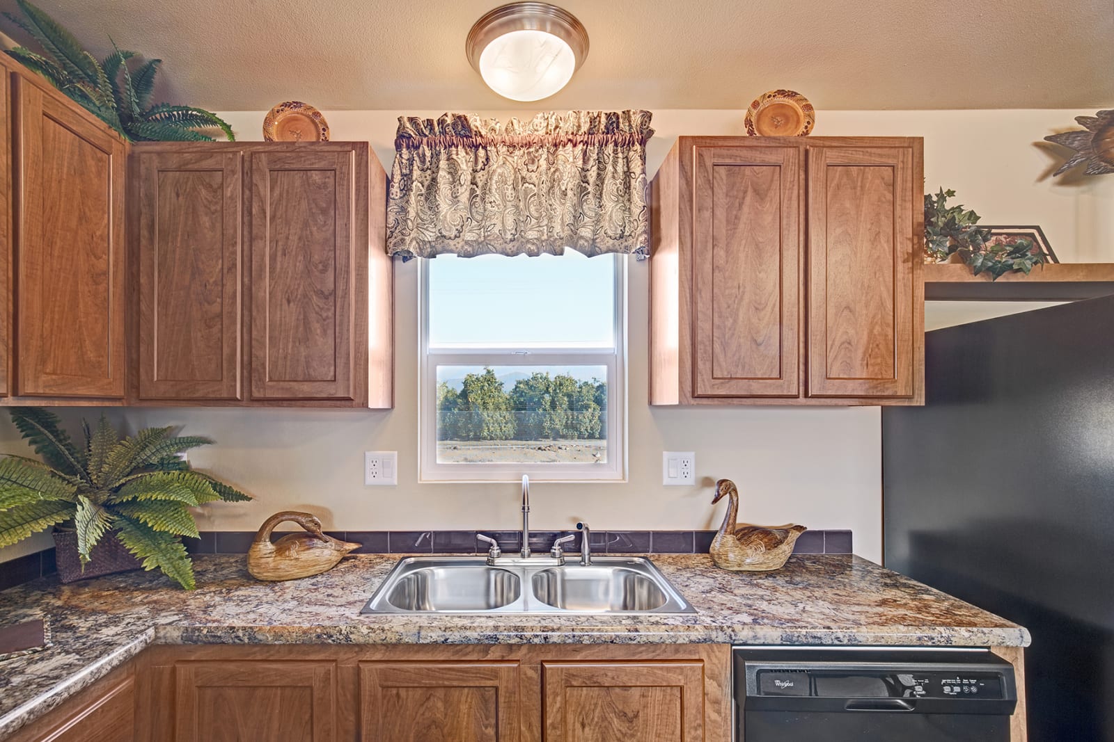 Kitchen with wooden cabinets, double sink, window with view of trees, and decorations.
