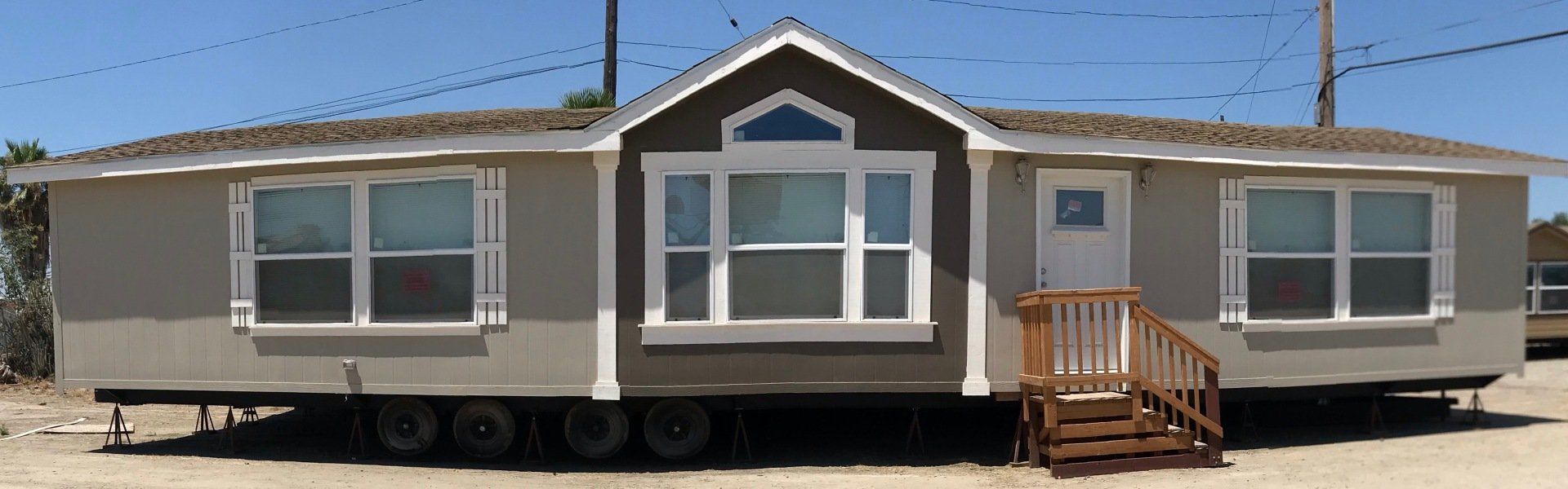 A manufactured home with light gray siding, white trim windows, and a brown front gable with a white door and wooden steps.