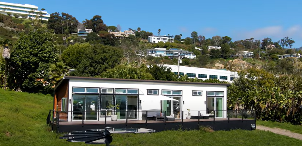 Modern, white house with deck overlooking a green hillside under a blue sky.