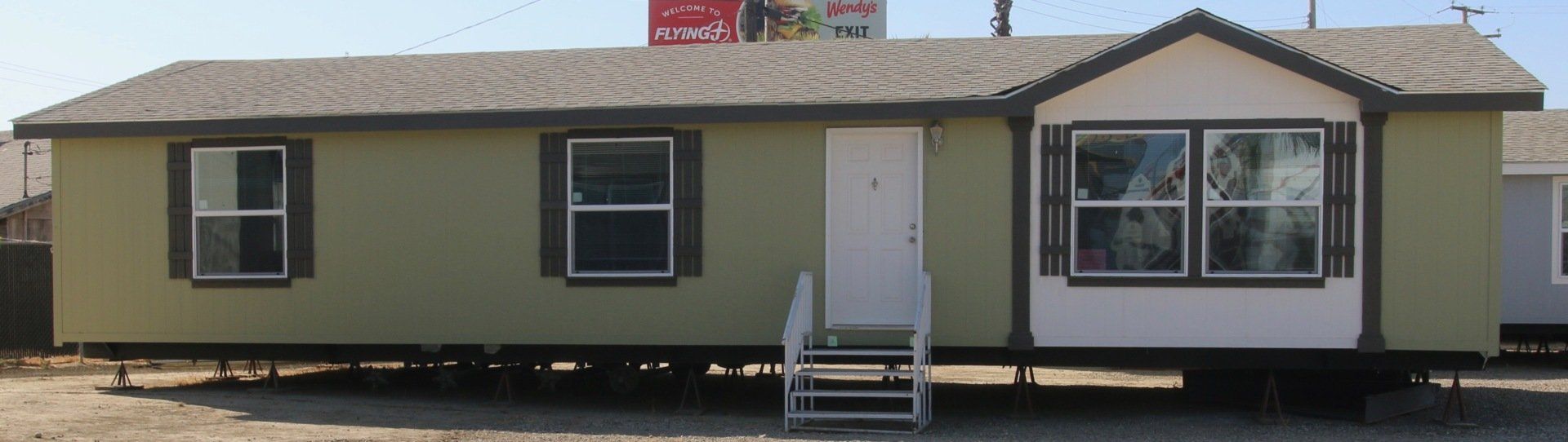 Green mobile home with white door and bay window, grey roof, and blue sky background.