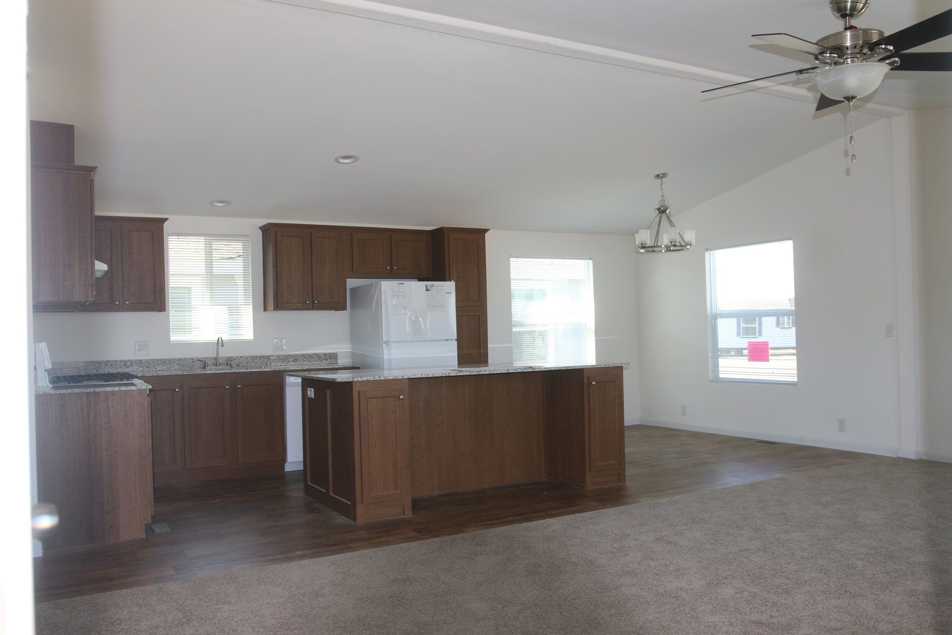 Kitchen with brown cabinets, island, and ceiling fan; windows and empty living space with brown carpet.
