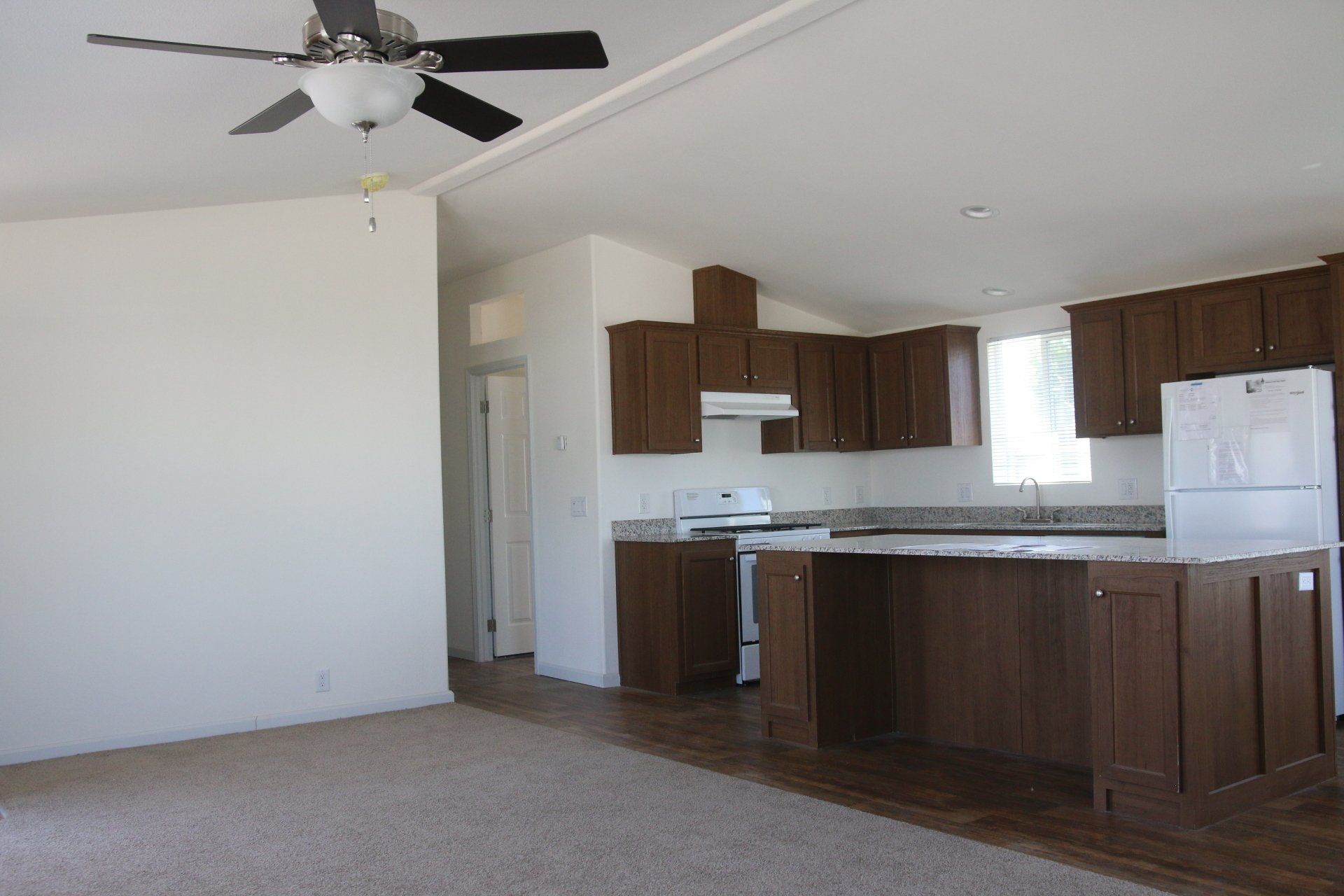 Open-concept living space with kitchen. Brown cabinets, white appliances, beige walls, and a ceiling fan.