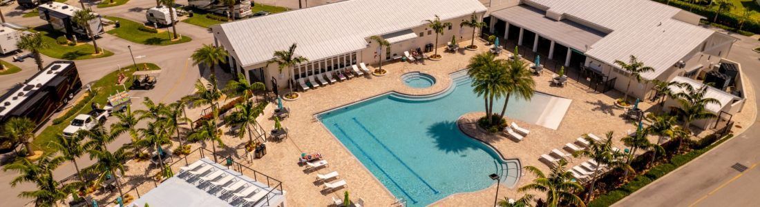 Aerial view of a resort with a large swimming pool, buildings, palm trees, and RVs.