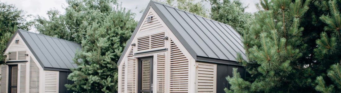 Two small wooden cabins with gray metal roofs nestled among green trees.
