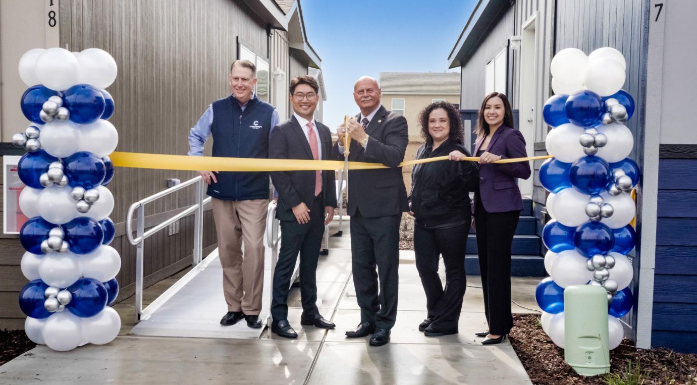 Ribbon-cutting ceremony outside a building, with five people holding scissors behind a gold ribbon and balloon columns.