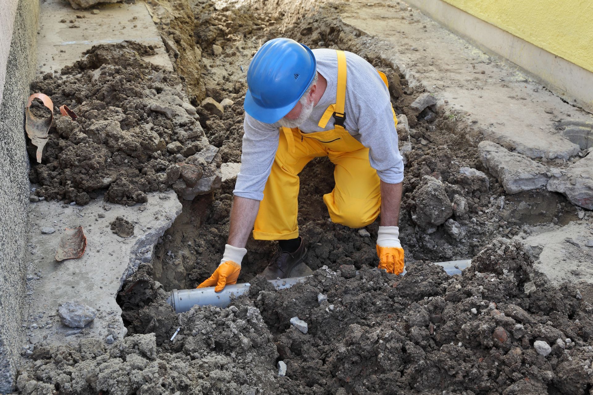 Construction worker in yellow overalls and blue hard hat shoveling dirt at a trench site