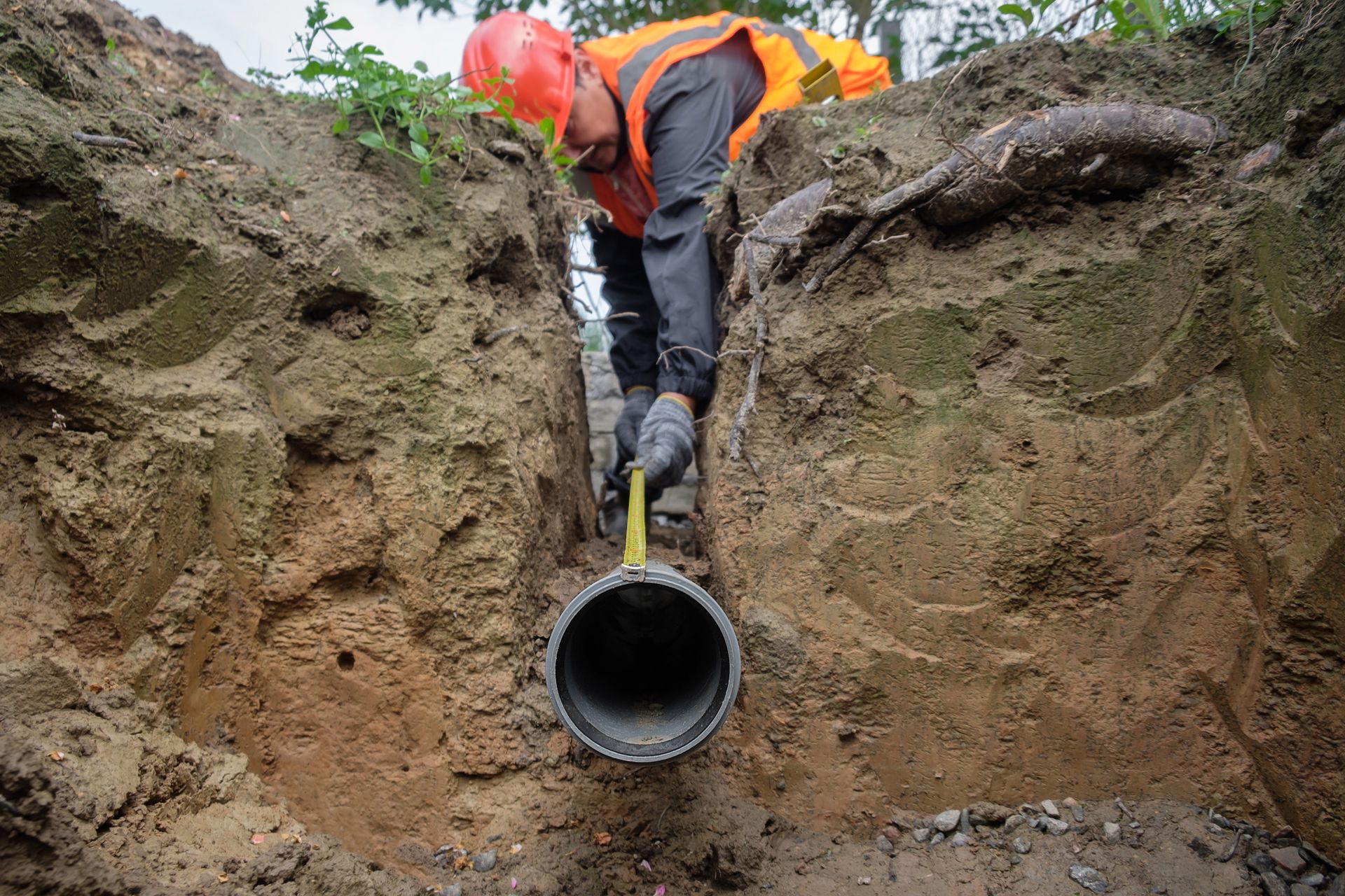 Worker in orange safety gear inspecting a large pipe in a muddy trench