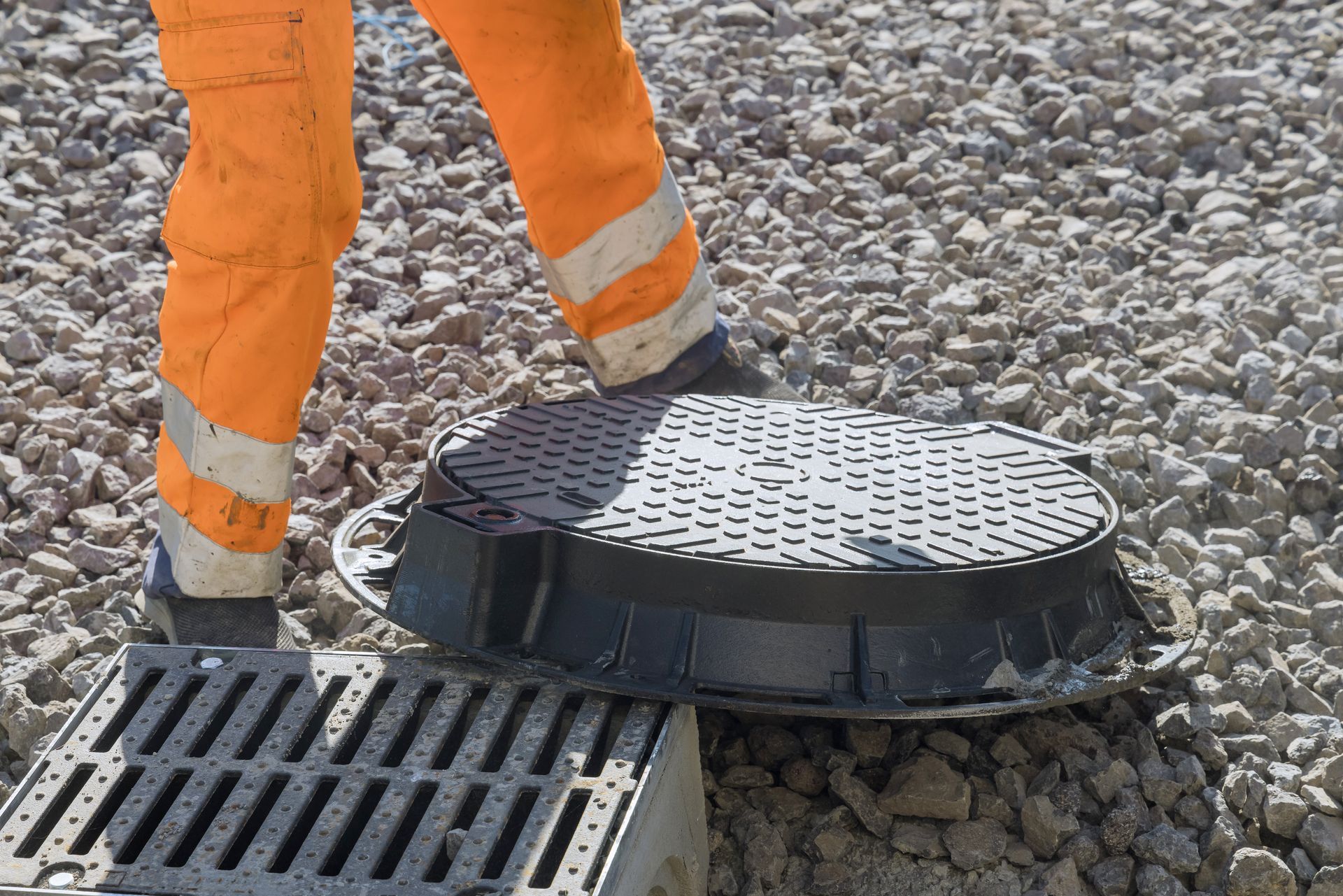 Worker in orange safety pants beside a round manhole cover and storm drain on gravel.