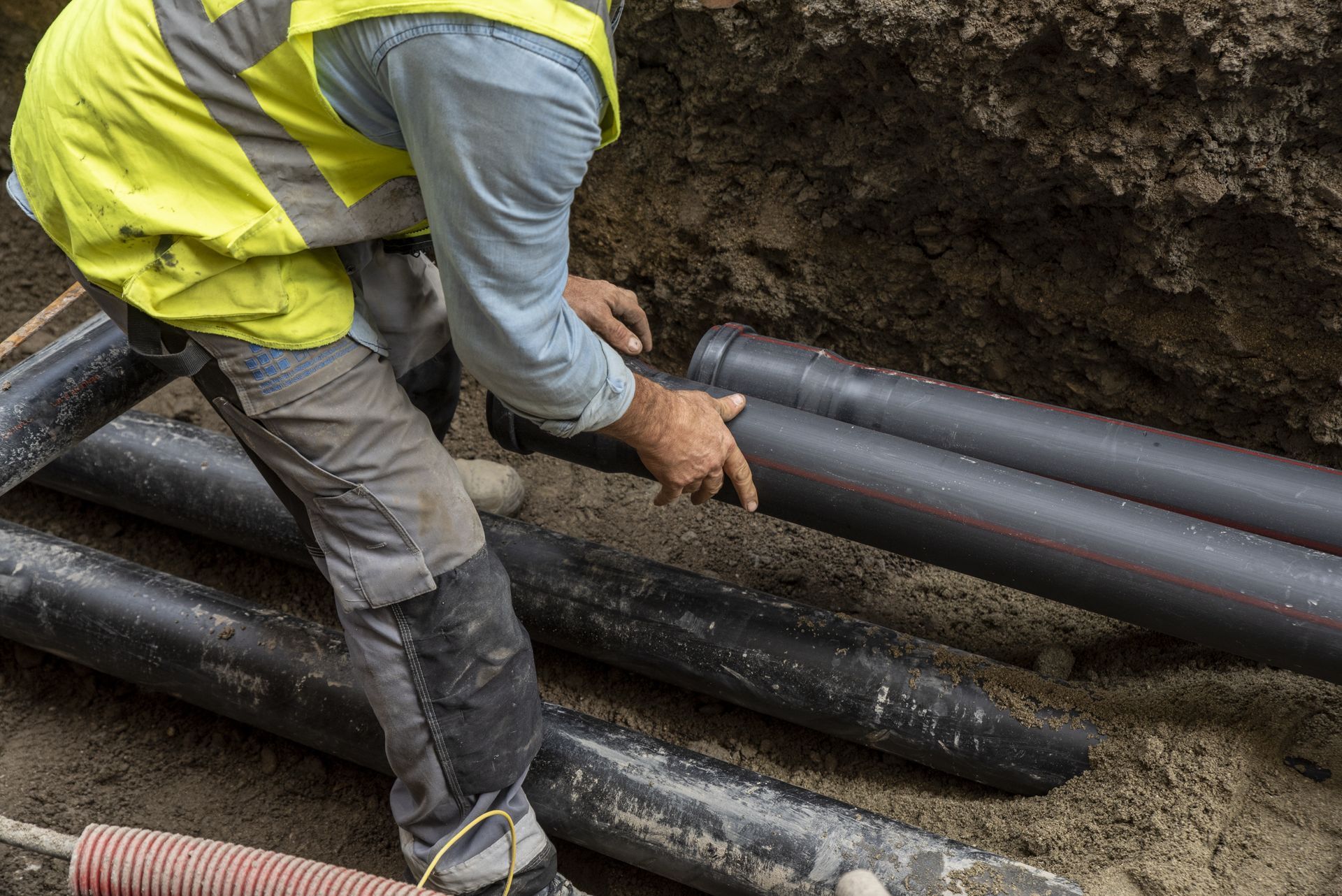 Worker in a yellow vest installs large black pipes in a muddy trench.